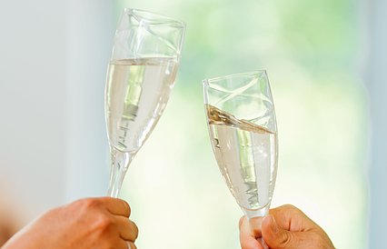 Zwei Hände halten mit Sekt gefüllte Gläser und stoßen an. Im Hintergrund verschwommen eine helle, freundliche Umgebung.